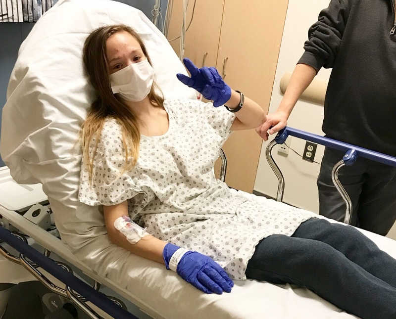 Woman with lupus and other issues lays in hospital bed holding up peace sign