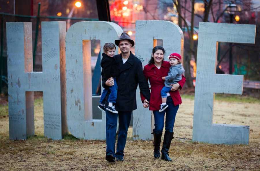 Husband and wife holds hands as they hold their two kids in front of large Hope sign