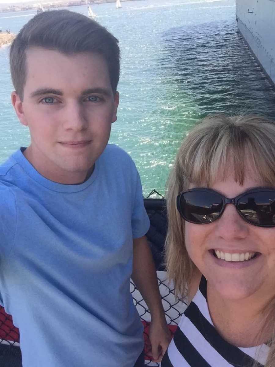 Woman smiles in selfie with son beside railing that looks out to body of water