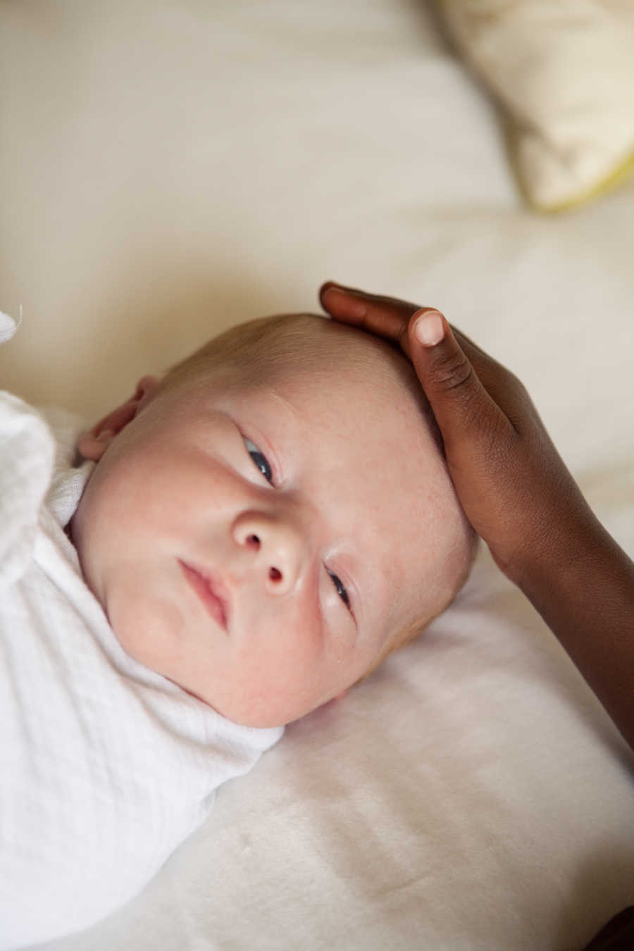 White baby lays swaddled in white blanket while African American child's hand rests on his head
