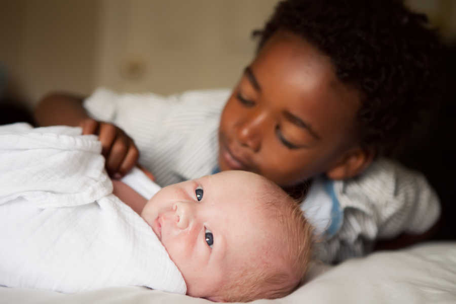 African American little boy lays next to white baby brother adjusting his blanket