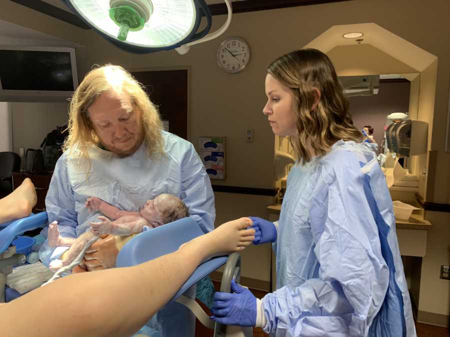 Doctor holds newborn baby in delivery room while adoptive mother stands beside him