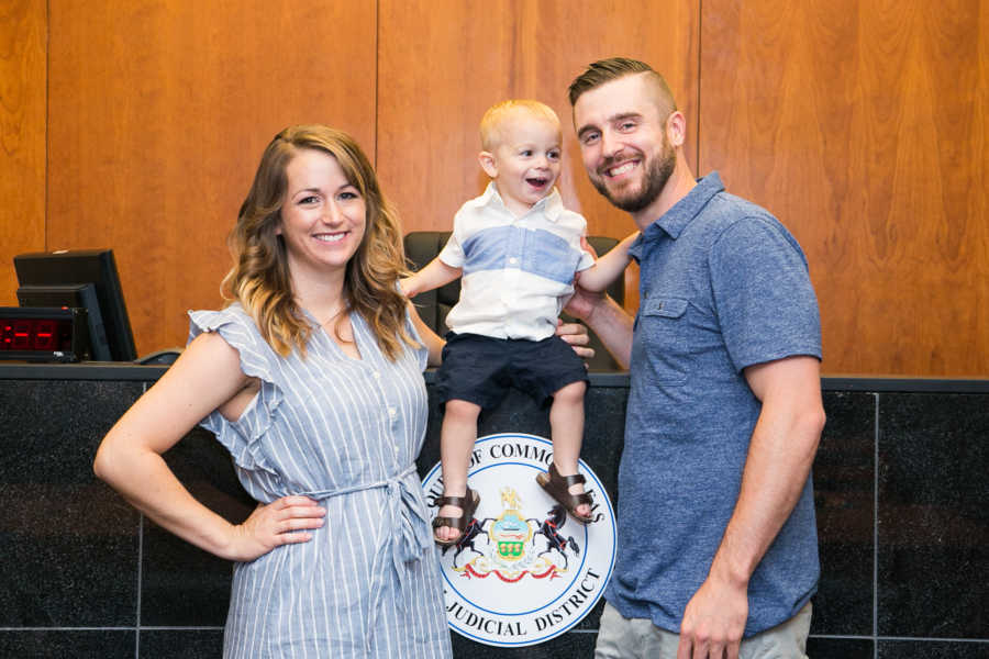 Husband and wife stand smiling in adoption court with their adopted son sitting on stand