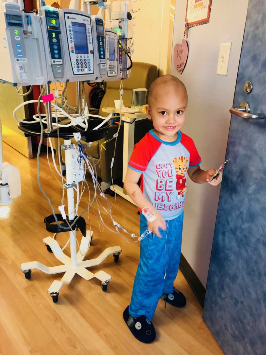 Little boy going through chemo stands smiling in hospital room hooked up to monitor