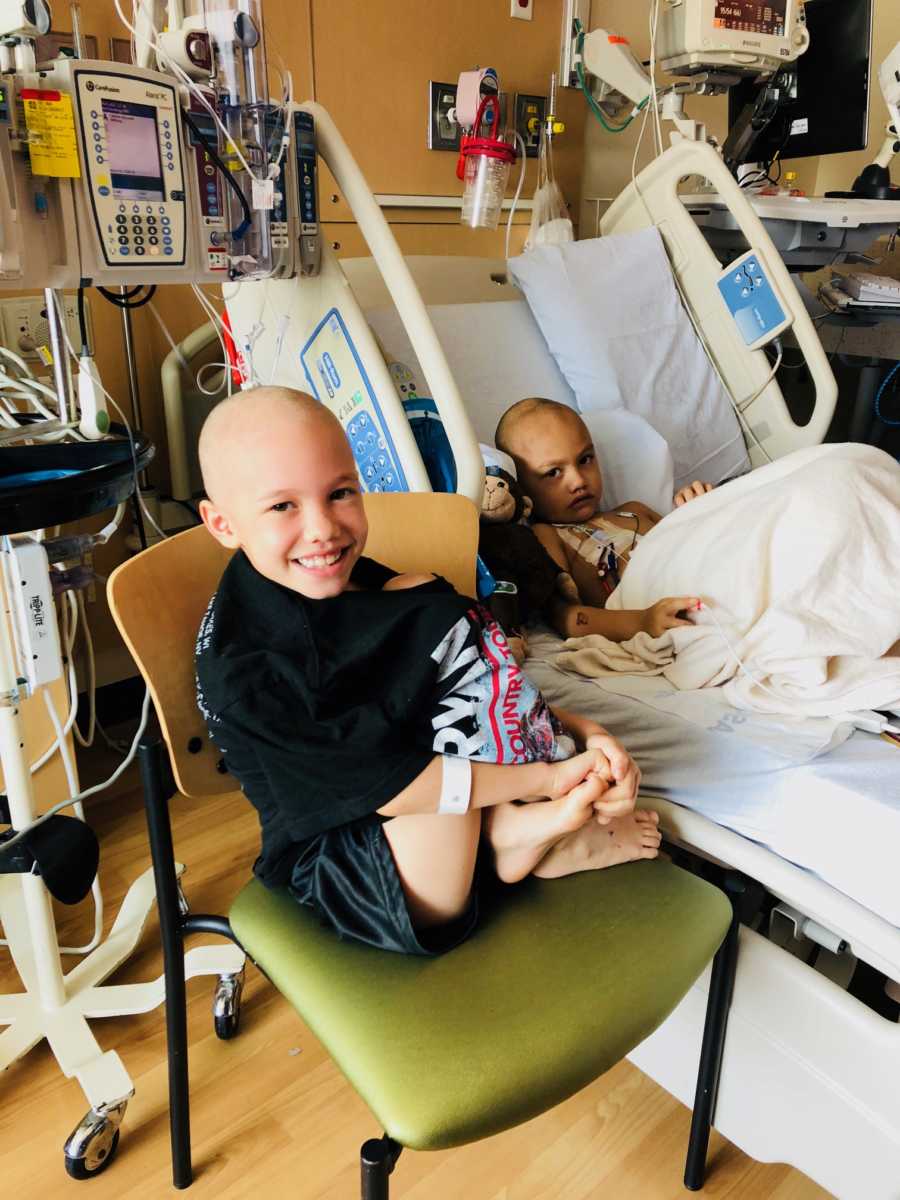 Sister with brain cancer sits in chair beside younger brother with brain cancer who sits hospital bed