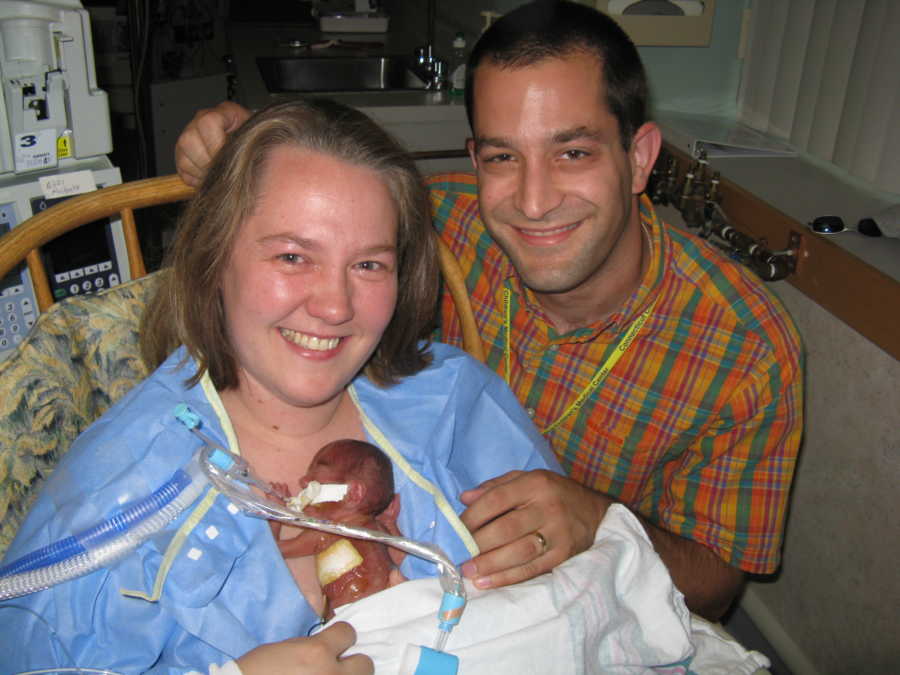 Mother sits in NICU with preemie baby laying on her chest while father smiles beside them