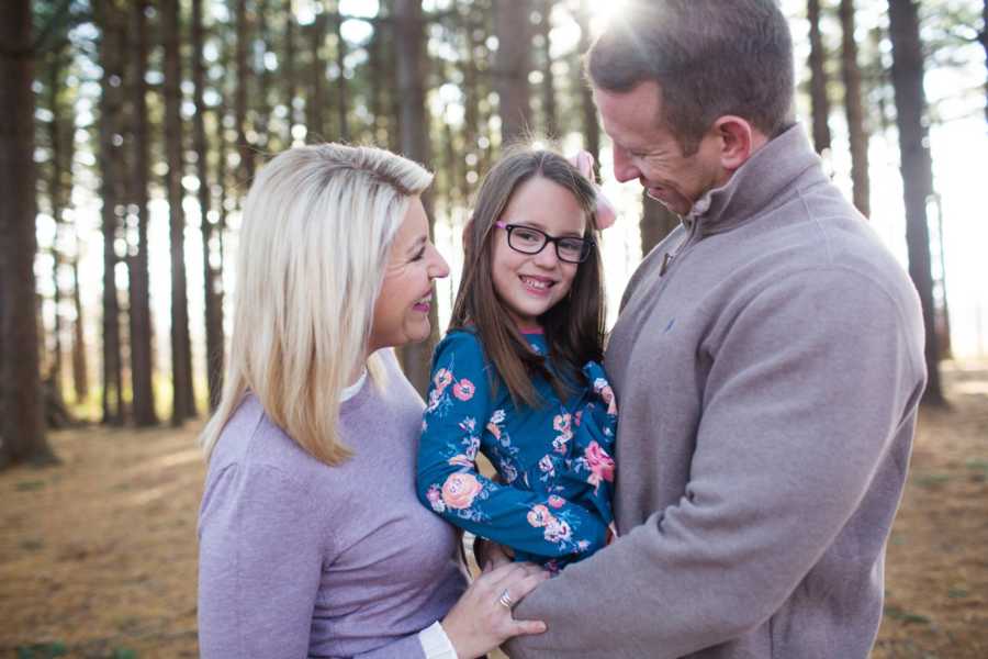 Father smiles as he holds young daughter in wooded area with wife beside him