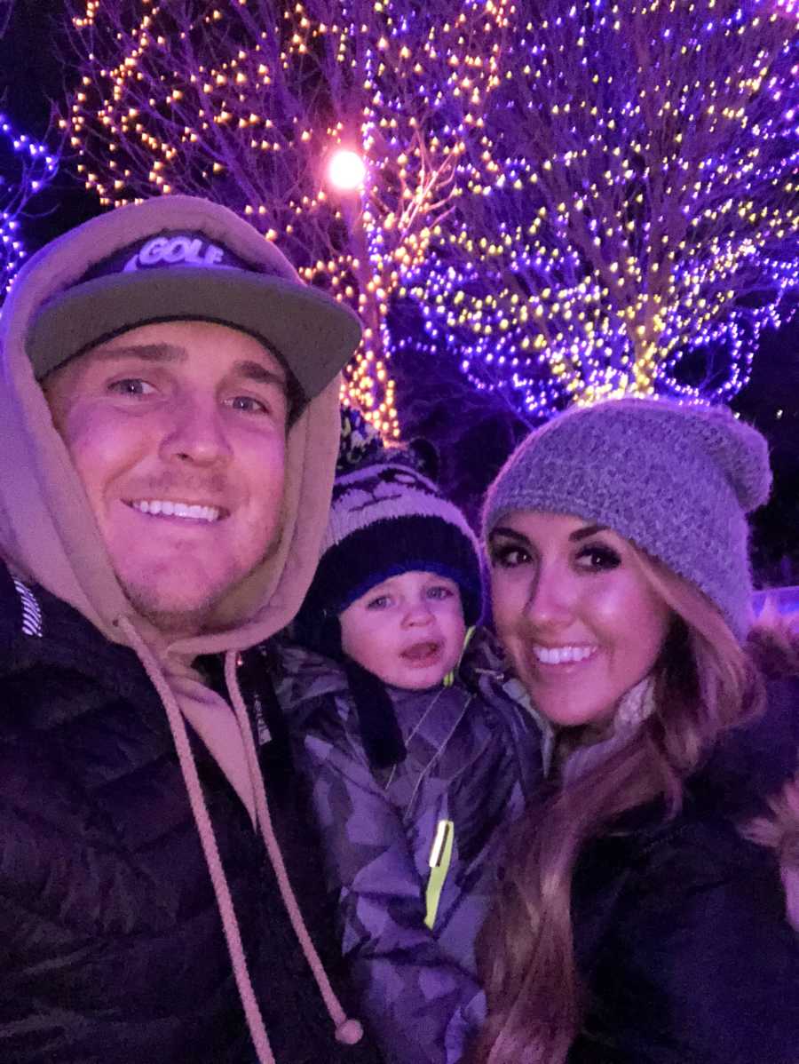 Husband and wife stand smiling in selfie with their nephew outside in front of trees wrapped in lights