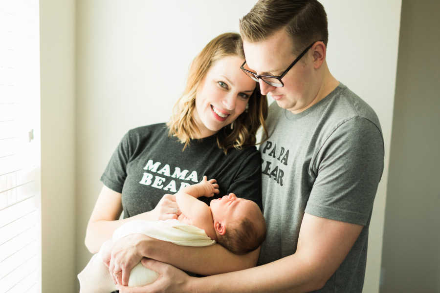 Husband stands behind wife as they hold their adopted newborn