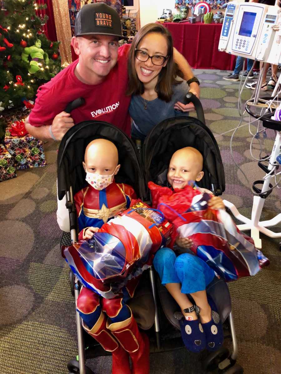Young siblings with brain cancer sit in stroller together while their parents stand smiling behind them