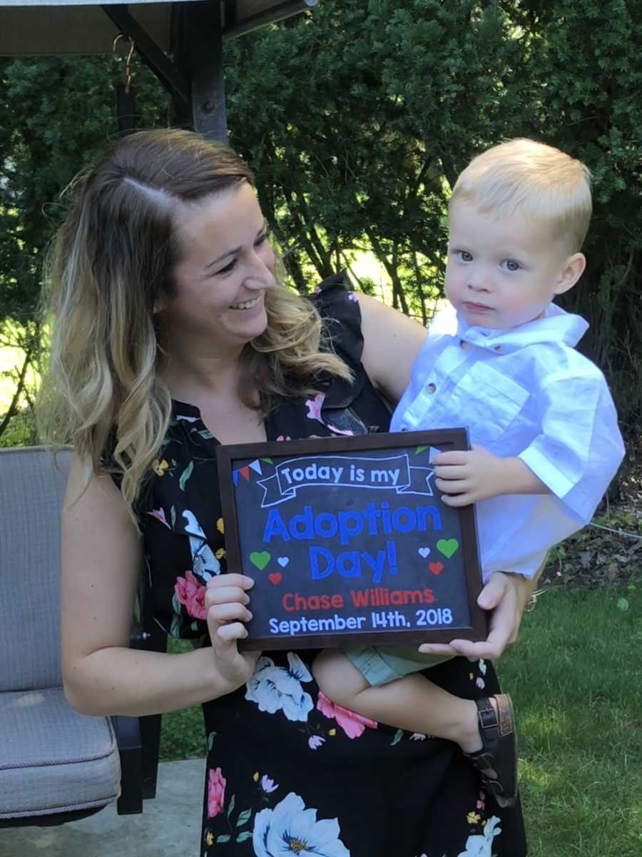 Woman stands outside holding sign announcing adoption of her foster son she holds