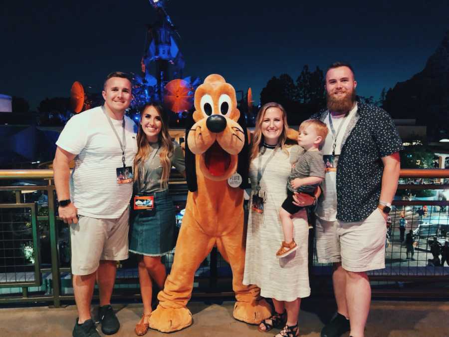 Husband and wife who are pregnant stand at Disney World beside Pluto, their nephew, wife's brother and sister-in-law