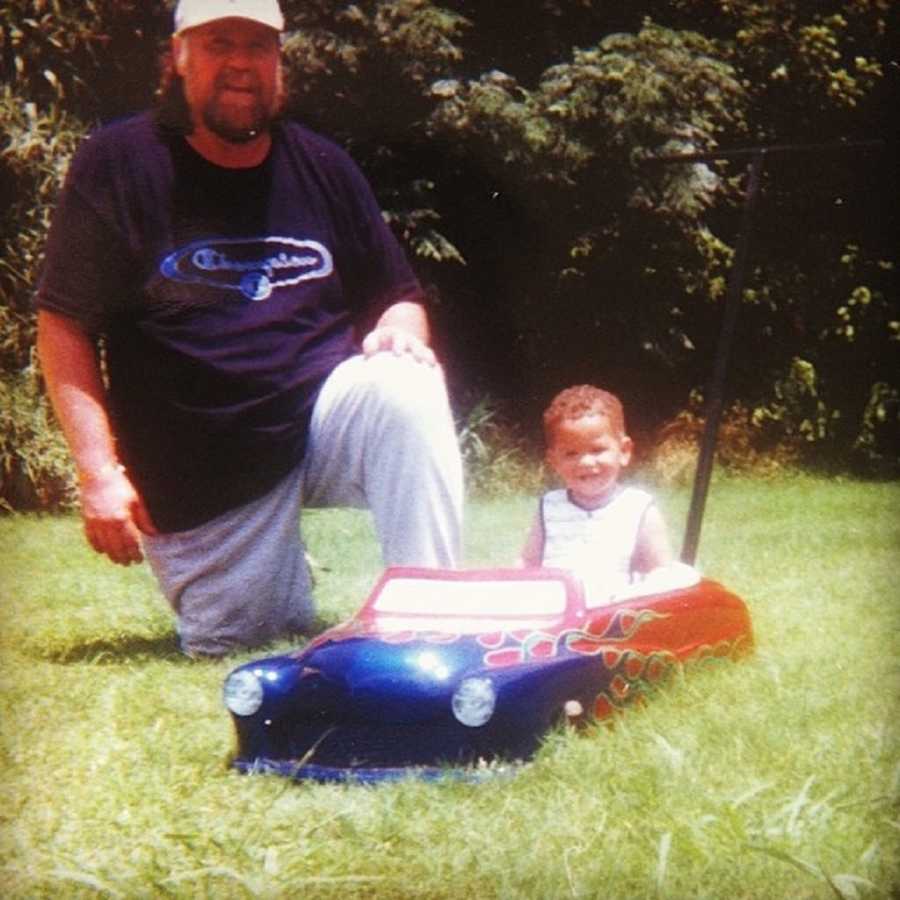 Man kneels on ground outside beside grandson who sits in toy car