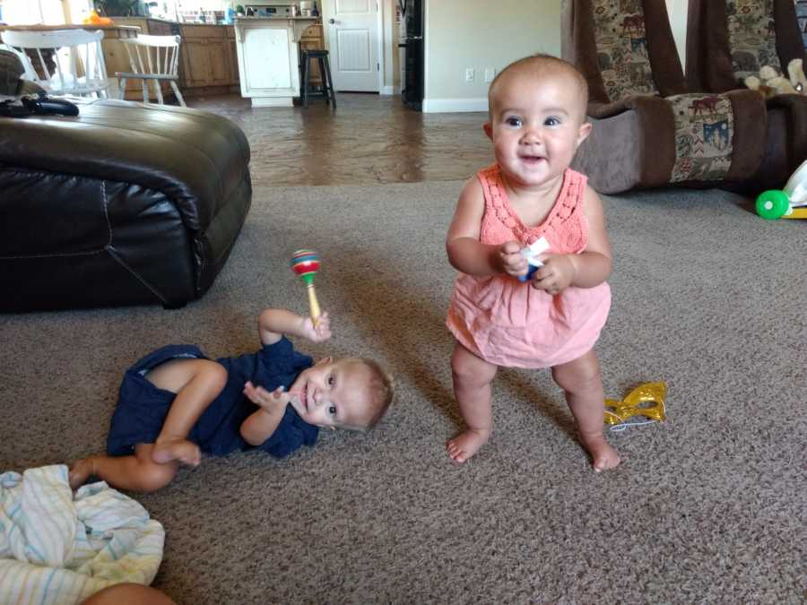 Baby with unknown disease lays on floor playing with toy while another baby stands beside him