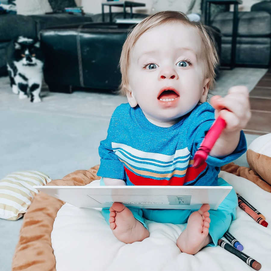 Little boy with dwarfism sits on floor holding onto Crayon