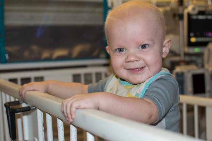 Baby stands smiling in hospital crib for chemo treatments