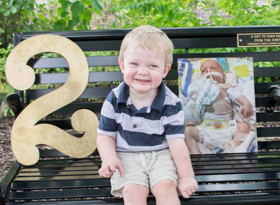 Little boy smiles while sitting on metal bench beside big number two and picture of him as baby in hospital