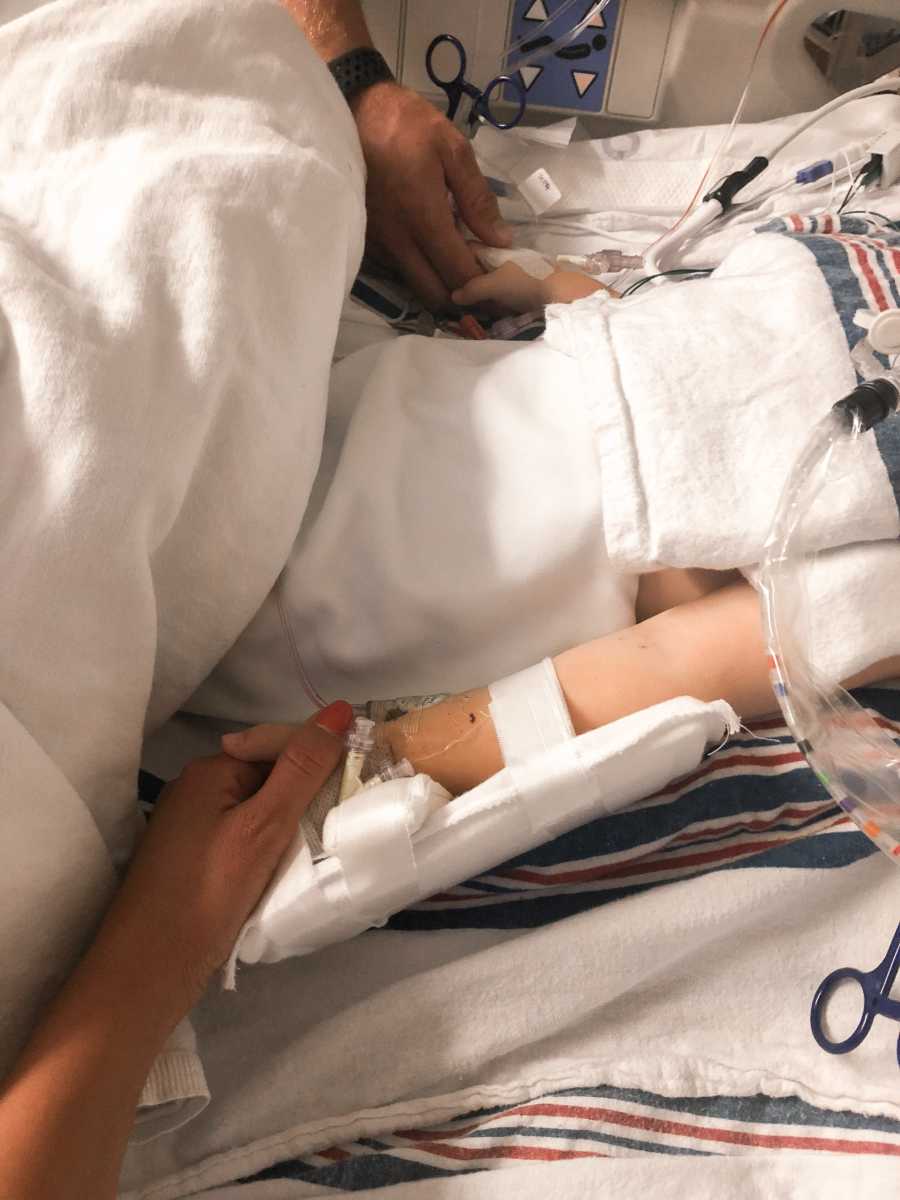 Aunt and uncle hold their nephew's hands who lays in hospital bed after having two heart attacks