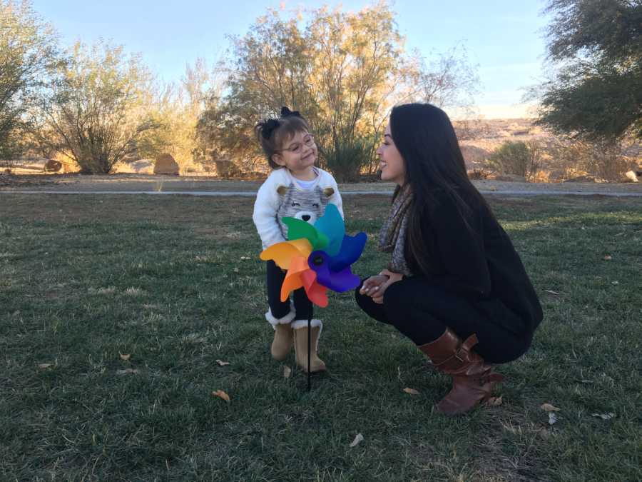 Mother crouches down beside daughter who stands behind rainbow pinwheel outside