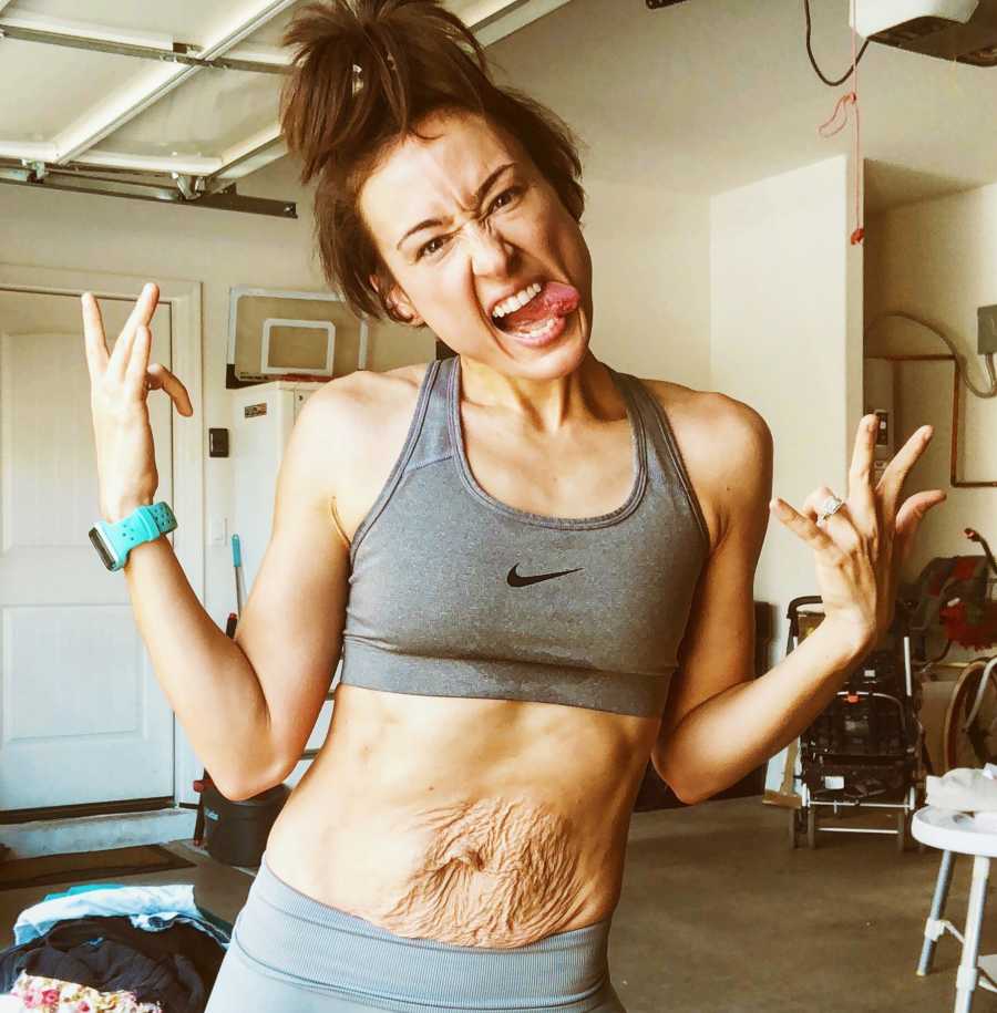 Mother with postpartum stomach stands in garage wearing sports bra and leggings sticking tongue out