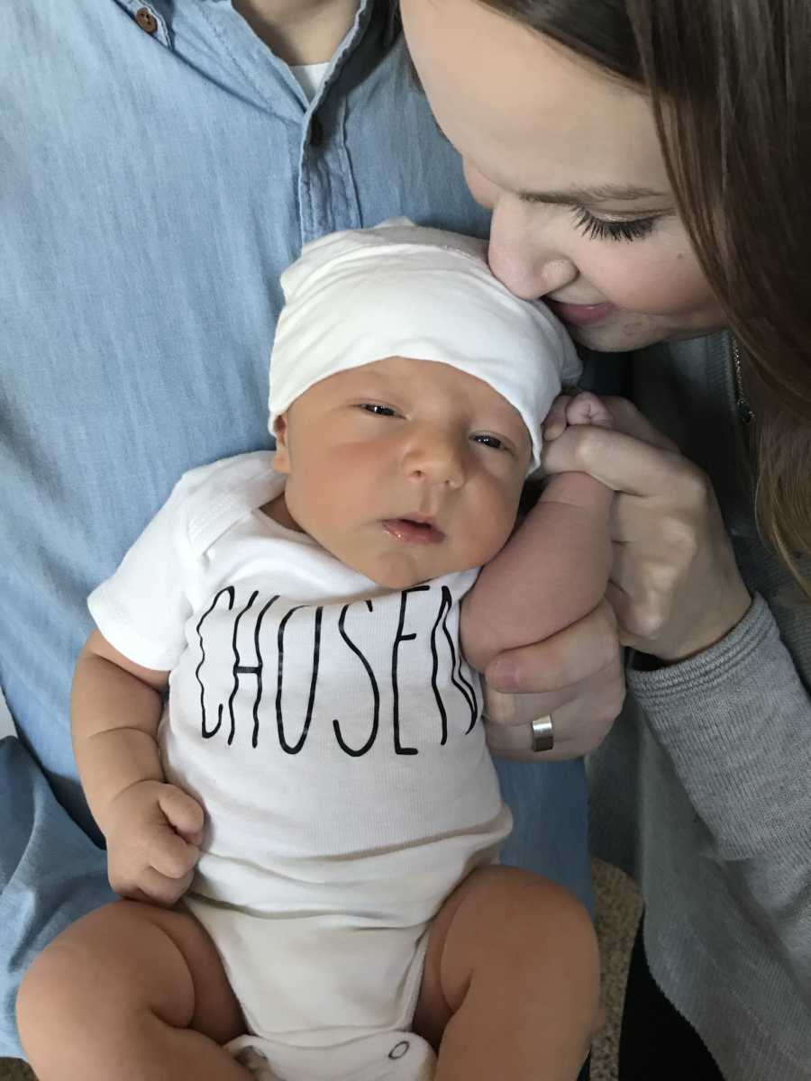 Adopted newborn wearing onesie that says, "Chosen" is being held by father as mother kisses his forehead