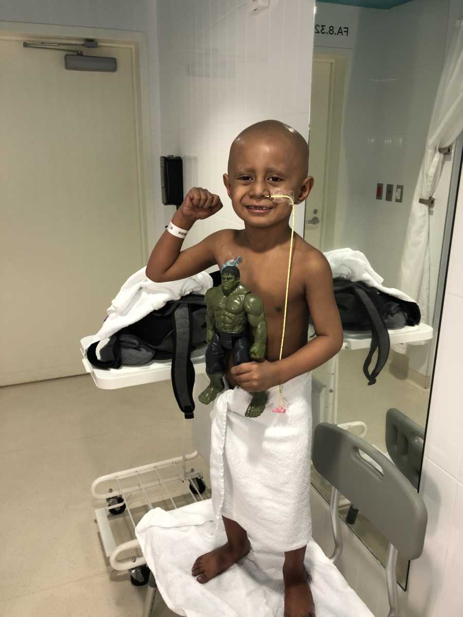 Little boy with cancer stands wearing only a towel while flexing muscles and holding toy Hulk
