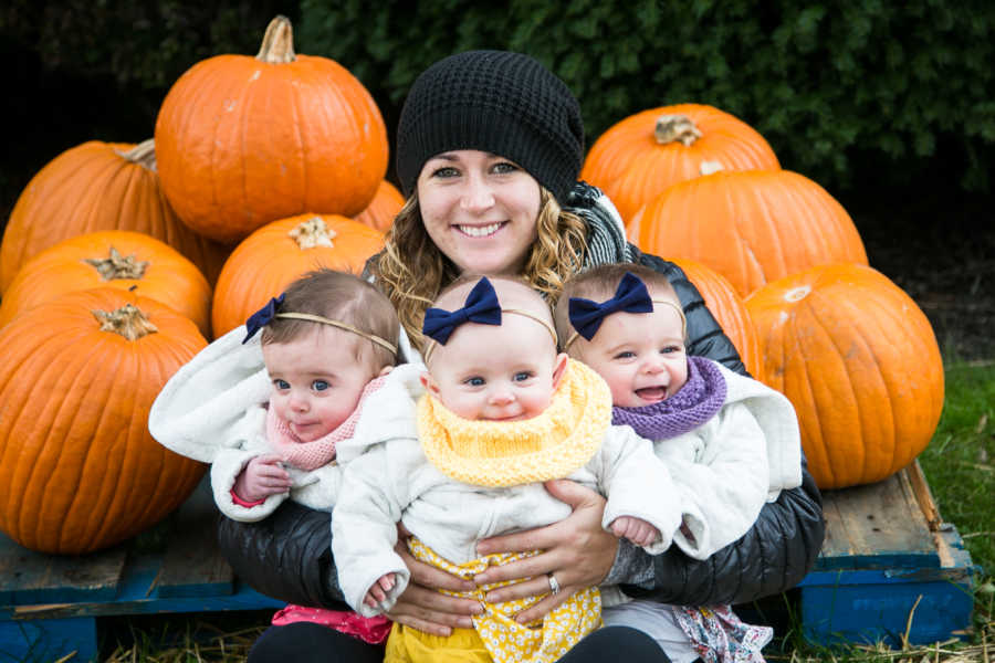 Mother sits outside holding triplets in front of pumpkins