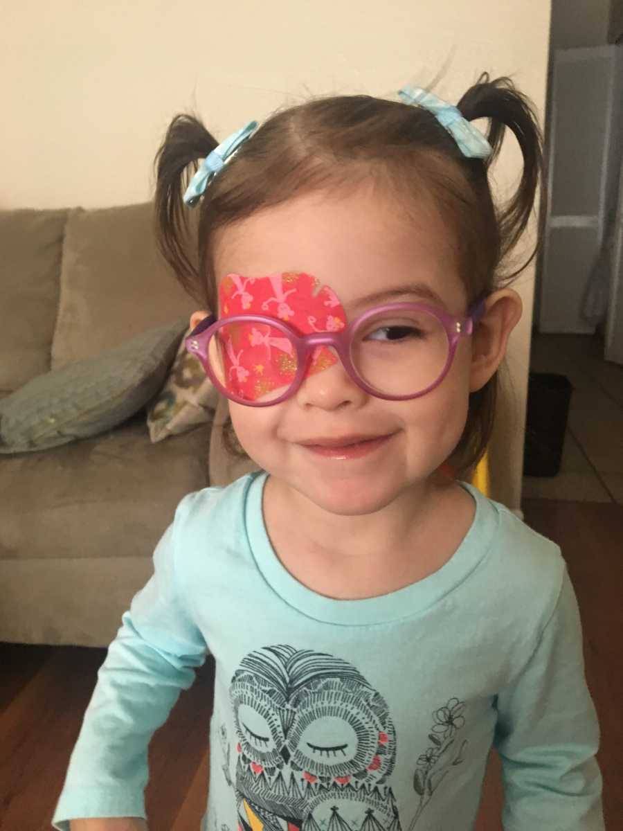 Little girl with strabismus stands smiling in home with pink glasses on and eye patch on left eye