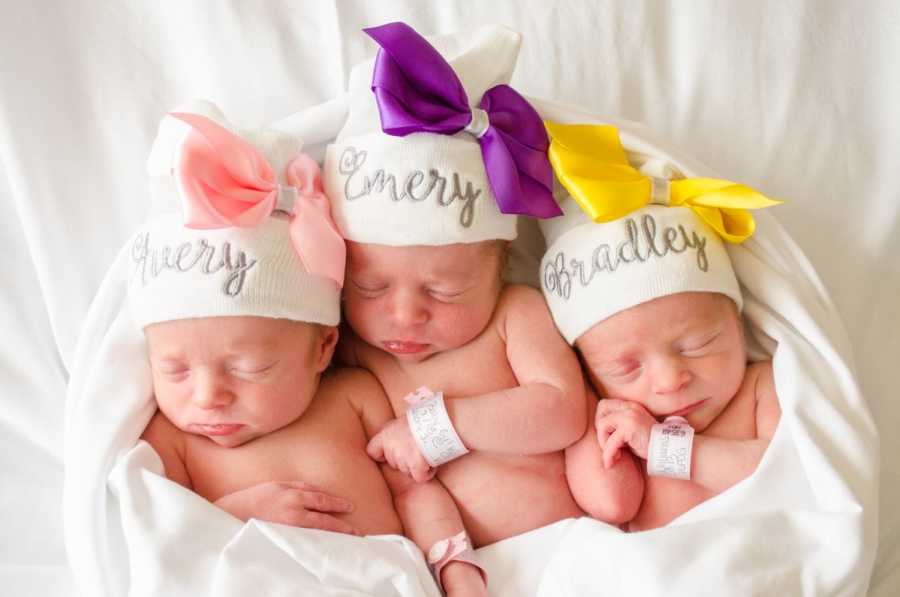 Triplets laying on their backs in white blanket with white hats with colored bows on them
