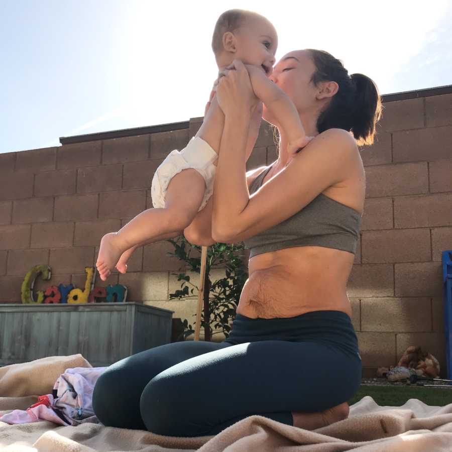 Mother with postpartum stomach sits on ground outside holding baby in air