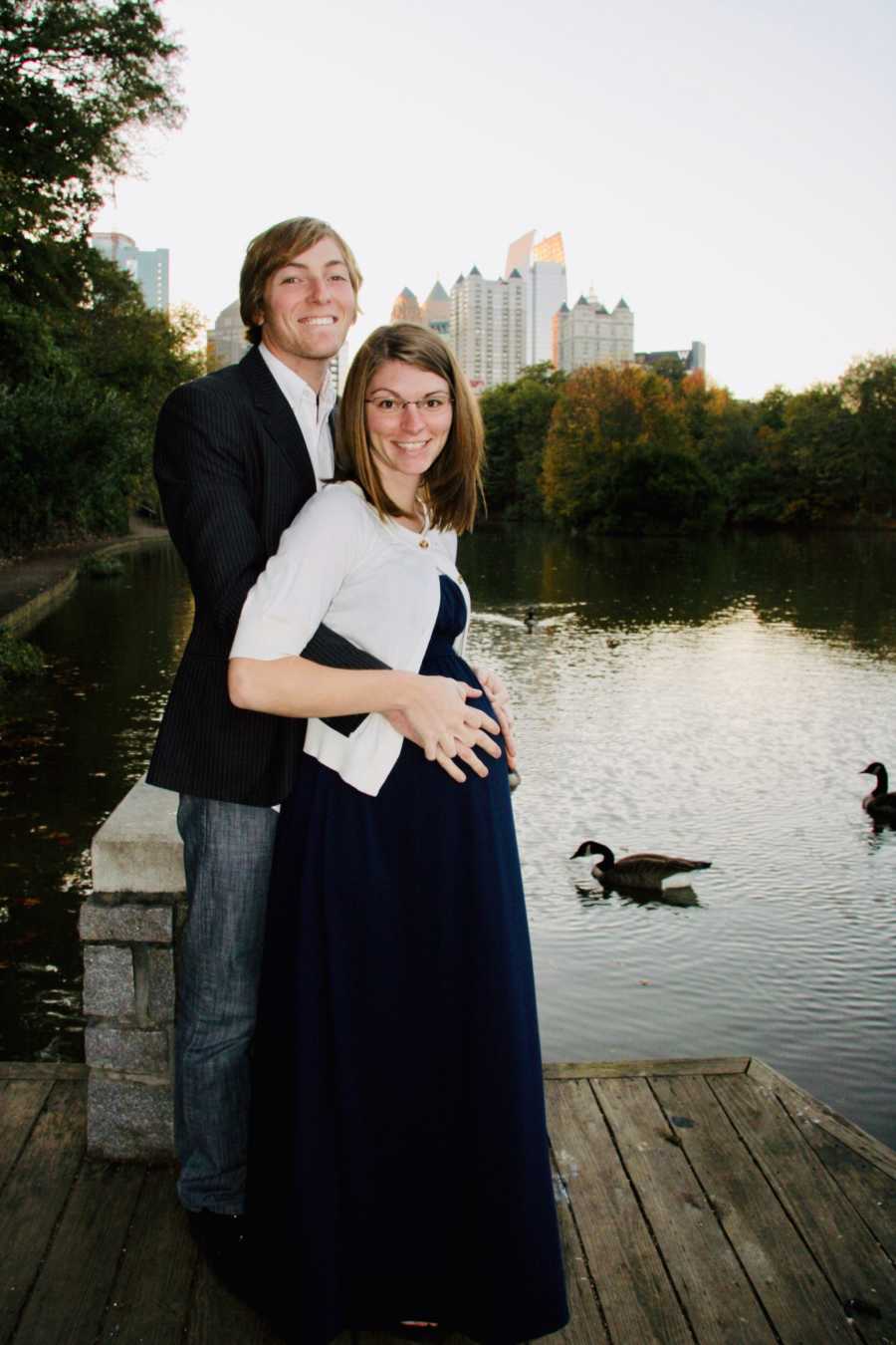 Husband stands behind holding pregnant wife on dock with city skyline in background