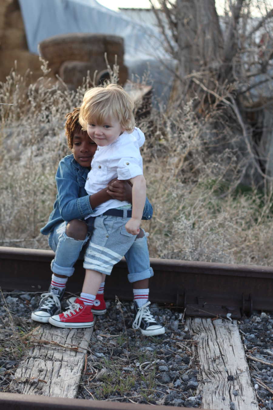 Young African American boy sits on train track hugging white little brother who stands in front of him