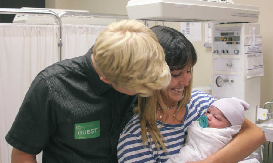 Woman smiles as she holds newborn and husband stands beside her