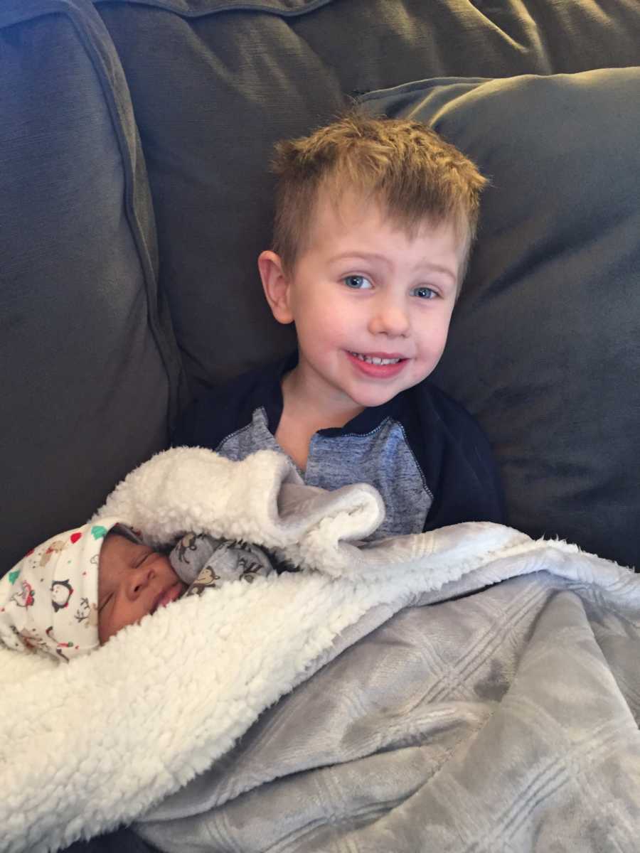 Little boy sits on couch with adopted baby brother sleeping in his lap