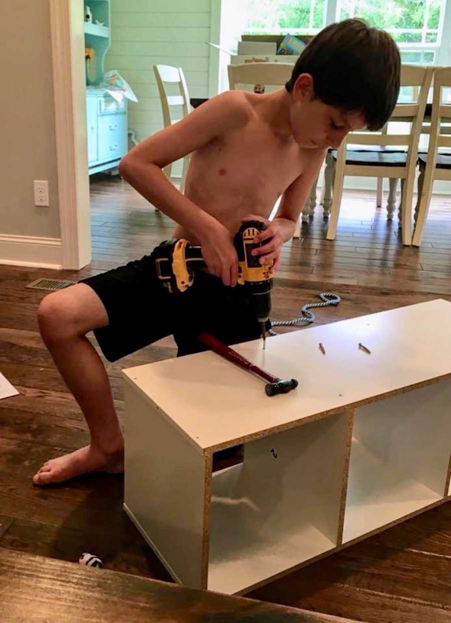 Little boy kneels on floor of home holding drill as he puts together book case