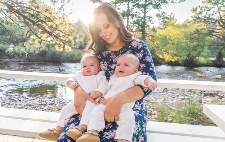 Mother sits smiling with two twin boys in her lap with body of water in background