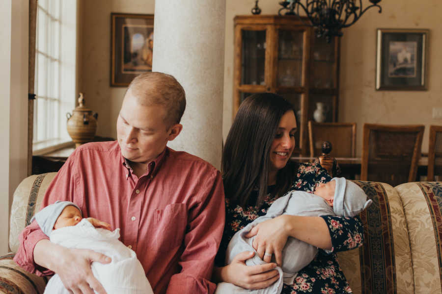 Husband and wife sit on couch at home each holding a twin boy