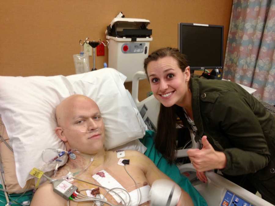 Man going through chemo lays in hospital bed as wife stands at his side with thumbs up