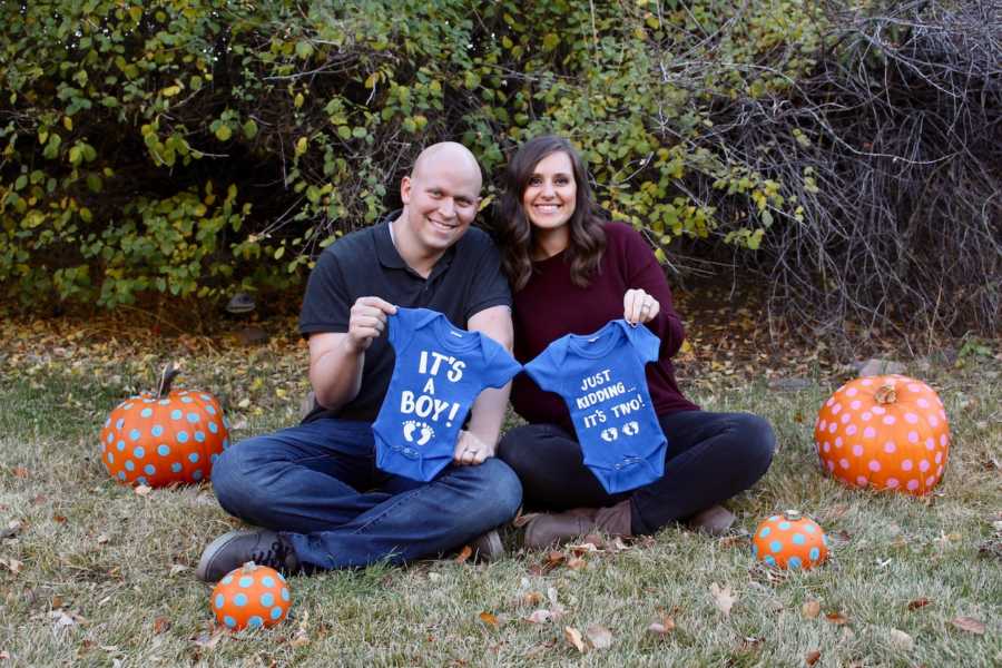 Husband who beat cancer sits on ground beside wife as they hold two onesies for their twin boys