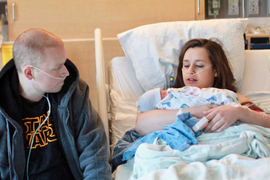 Woman lays in hospital bed holding her newborn as her husband with cancer sits beside her