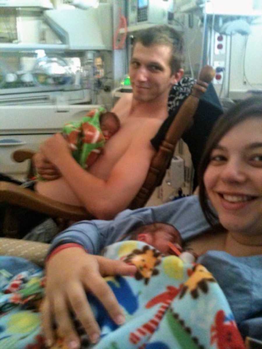 Mother smiles as she holds newborn to her chest in selfie while husband holds other twin 
