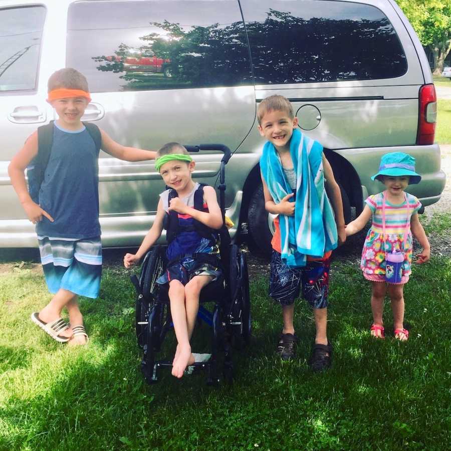 Boy with cerebral palsy sits in wheel chair in yard beside twin and two other siblings