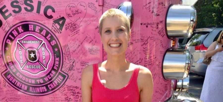 Woman with cancer stands smiling in front of pink truck