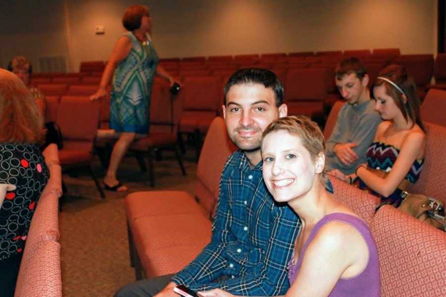 Husband sits smiling beside wife who has cancer and has since passed in room with rows of chairs