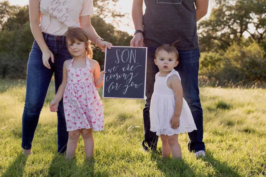 Little girls stand outside holding sign announcing adoption of brother with parents standing behind them