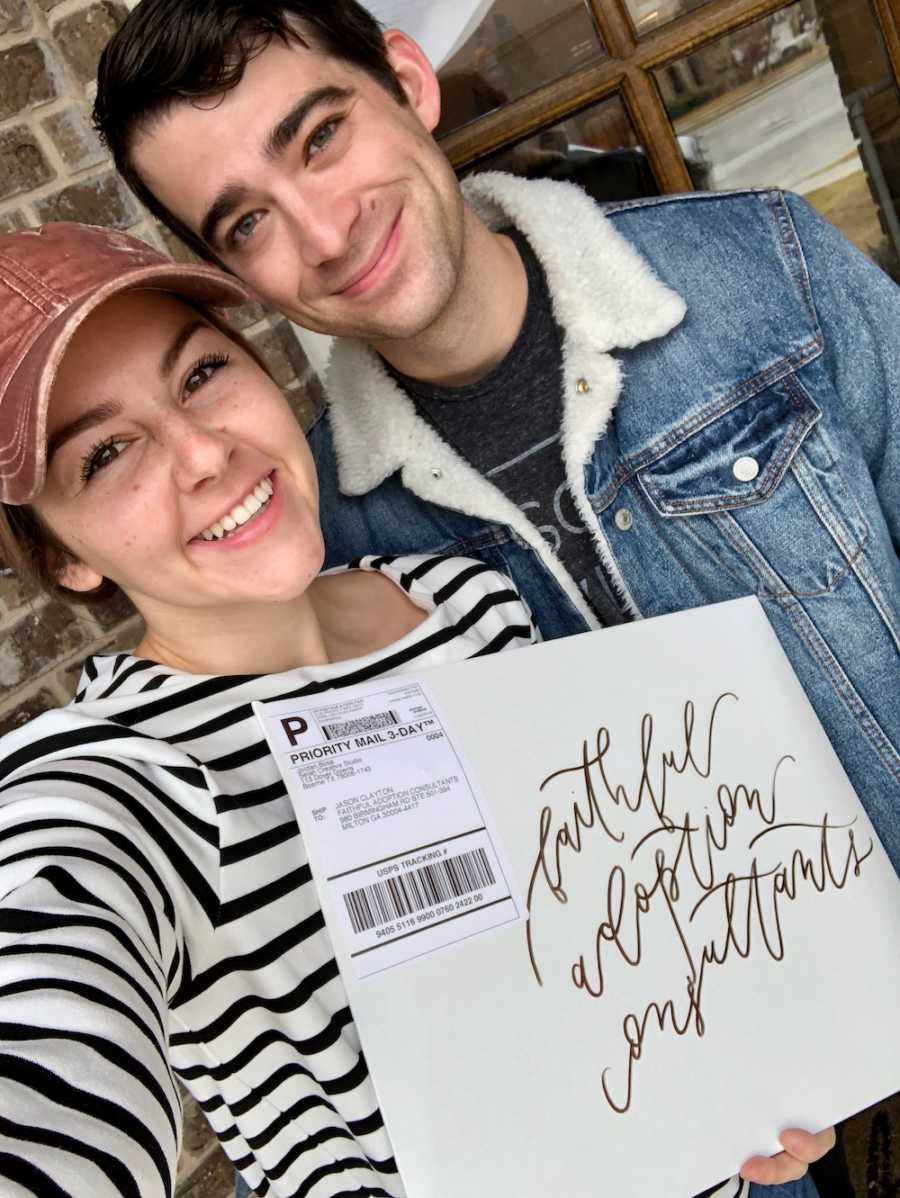 Husband and wife stand outside home holding packet of mail that contains adoption paperwork