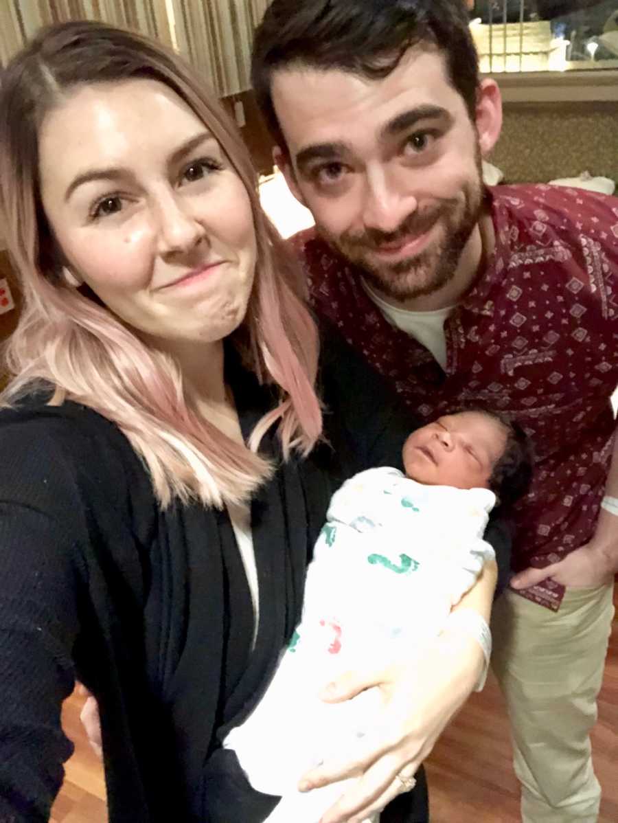 Woman smiles in selfie with newborn in her arms and husband beside her