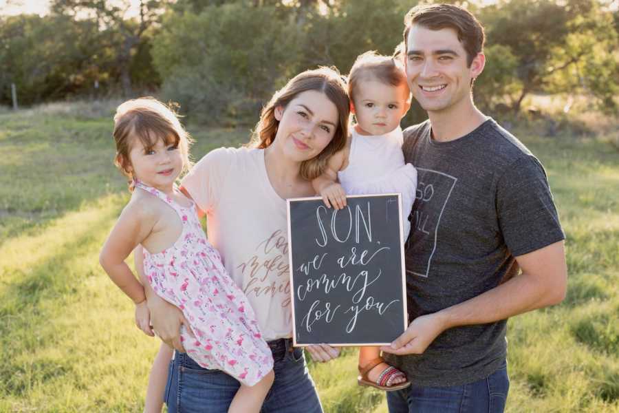 Husband and wife stands outside with their two daughters as they hold sign that says, "son we are coming for you"