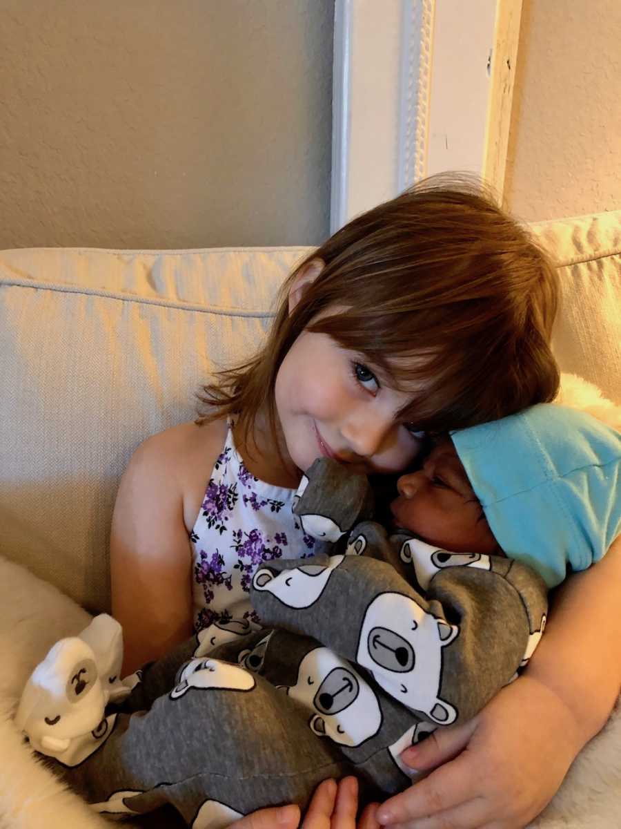 Little girl sits on couch with adopted newborn brother in her arms