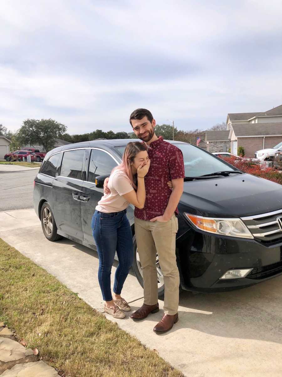 Wife holds hand over her mouth beside husband in driveway who are going to adopt a child 